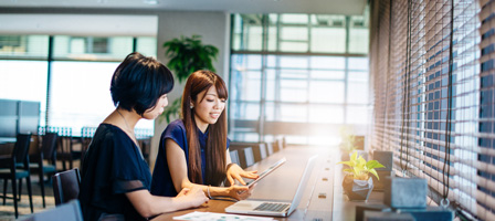 two women sitting at a table looking at a ipad with a laptop in front of them