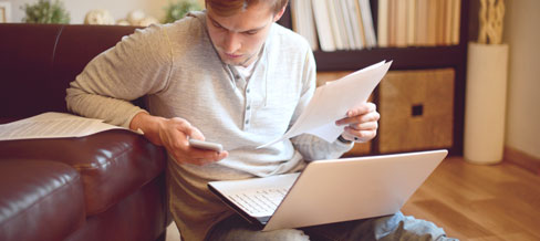 a man looking at his phone with paperwork in his hand and a laptop in his lap